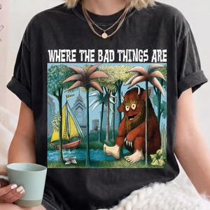 Può includere: Una t-shirt grigio antracite a maniche corte con una stampa grafica che riporta il testo "WHERE THE BAD THINGS ARE" sopra l'illustrazione di un mostro marrone peloso seduto in una foresta vicino a una barca a vela con una città sullo sfondo.