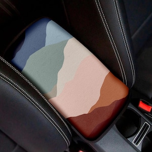 May include: A colorful abstract design car armrest cover with a geometric pattern of blue, green, pink, and brown. The cover is made of a soft, textured fabric and fits over the armrest of a car seat.