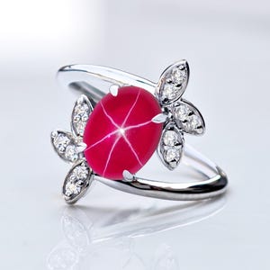 May include: A silver ring with a striking red star ruby gemstone. The oval ruby has a six-ray star pattern. The ring is embellished with small, clear gemstones arranged in a floral pattern on either side of the ruby.
