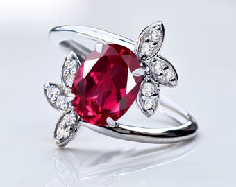 Puede incluir: Anillo de plata con una gran gema de rubí ovalada. El anillo tiene un diseño de banda dividida con dos acentos florales a cada lado del rubí, cada uno adornado con pequeños diamantes redondos. El anillo se muestra sobre un fondo blanco.