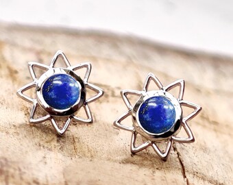 Könnte beinhalten: Ein Paar silberfarbene Ohrstecker. Jeder Ohrring hat einen blauen Edelstein in der Mitte, umgeben von einem sternförmigen Design. Die Ohrringe sind auf einer Holzoberfläche platziert, die ihre Details und Farbe hervorhebt.