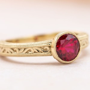 Puede incluir: Un anillo de oro con una piedra preciosa roja engastada en un engaste de bisel. La banda está grabada con un delicado diseño floral.