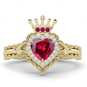 Puede incluir: Un anillo de oro con un rubí en forma de corazón rodeado de diamantes. El anillo tiene un diseño de corona con tres rubíes en la parte superior.