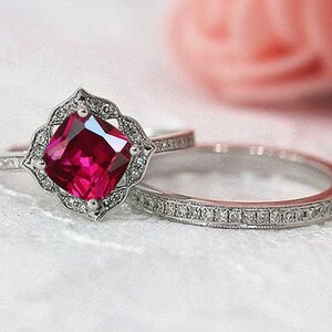 May include: A silver ring set with a large, square-shaped ruby gemstone surrounded by smaller diamonds. The ring is set on a white background.