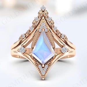 Peut inclure: Bague en or rose avec un design géométrique. La bague présente une pierre centrale irisée en forme de cerf-volant, entourée de petites pierres précieuses claires. La bande a un motif décoratif complexe. Une bande assortie se trouve en dessous.
