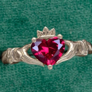 May include: A red heart-shaped gemstone set in a gold Claddagh ring. The ring features two hands clasping a heart with a crown on top.