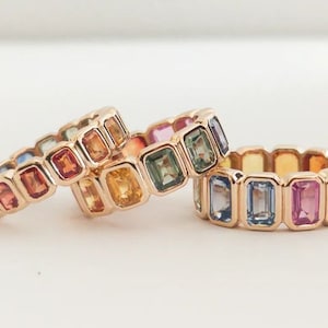 May include: Three gold rings set with colourful gemstones. Each ring features a row of rectangular-shaped gemstones in a rainbow colour pattern.