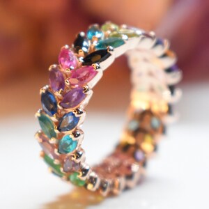 May include: A gold ring with a band of colorful gemstones. The stones are arranged in a row and are set in a leaf-like pattern. The ring is a beautiful and unique piece of jewelry.