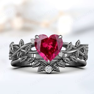May include: A black metal ring set featuring a heart-shaped, ruby-red gemstone as the centerpiece. The ring is adorned with leaf and vine detailing, and small, clear accent stones. The set includes a matching band with similar leaf and vine designs.