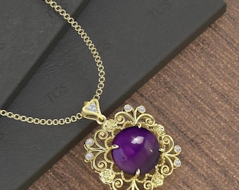 May include: A gold-colored pendant necklace featuring a large, round, deep purple gemstone. The gemstone is set in a decorative, square-shaped frame with small, clear accent stones. The necklace chain is also gold-colored.