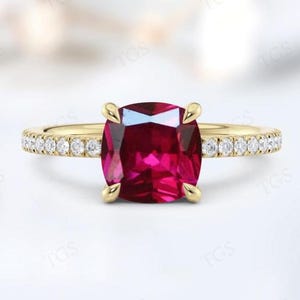 Pode incluir: Um anel de noivado de ouro com uma pedra de rubi de corte almofada e uma banda de diamantes.