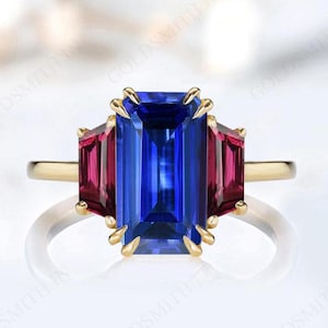 Puede incluir: Un anillo de oro con una gran piedra preciosa azul de talla esmeralda, flanqueada por dos piedras preciosas rojas de talla esmeralda más pequeñas.