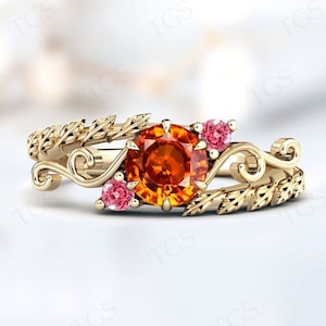 Puede incluir: Un anillo de oro con una gran piedra preciosa naranja y dos piedras preciosas rosas más pequeñas. El anillo está diseñado con una rama frondosa y un delicado remolino.