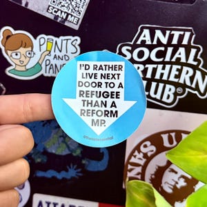 Puede incluir: Una pegatina redonda azul claro con el texto blanco "I'D RATHER LIVE NEXT DOOR TO A REFUGEE THAN A REFORM MP." y una flecha blanca que apunta hacia abajo. Otras pegatinas son visibles en el fondo.