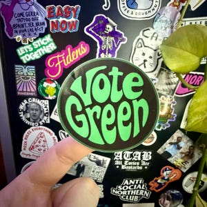 Puede incluir: Una pegatina redonda con la frase "Vote Green" en verde brillante y una fuente retro sobre un fondo negro. La pegatina está sujeta por un dedo, con otras pegatinas visibles en el fondo.