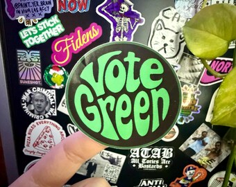 May include: A round sticker with the words "Vote Green" in bright green, groovy font on a black background. The sticker is held by a finger, with other stickers visible in the background.