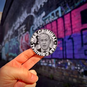 Puede incluir: Una pegatina en blanco y negro con una foto de un hombre y el texto "WAR CRIMINAL WAR CRIMINAL" alrededor del borde.