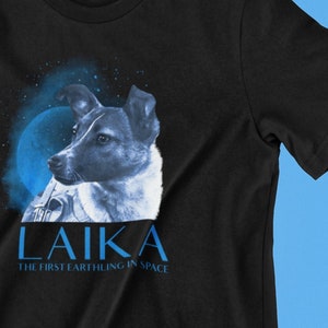 以下が含まれることがあります： 宇宙で最初の犬、ライカの青と白のイラストが描かれた黒いTシャツ。イラストの下には「ライカ」と「宇宙で最初の地球人」という文字が青で印刷されています。