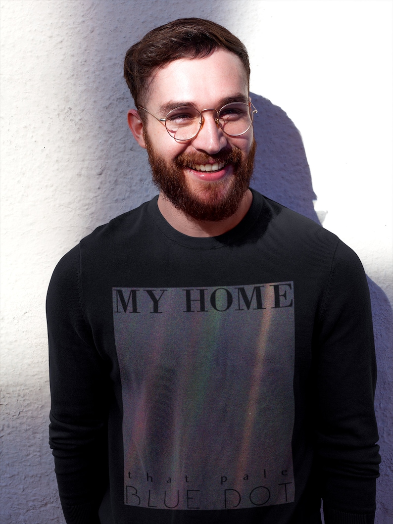 Puede incluir: Una camiseta negra de manga larga con un estampado gr&aacute;fico de una nebulosa colorida con el texto "MY HOME that pale BLUE DOT" en la parte delantera.