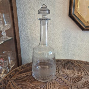 Könnte beinhalten: Eine klare Glas-Dekantierkaraffe mit einem Verschluss. Die Karaffe hat einen langen Hals und einen bauchigen Boden mit einem dekorativen geätzten Muster. Der Verschluss hat eine runde Form mit einem kleinen Knopf oben. Die Karaffe steht auf einem Holztisch.