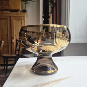 Könnte beinhalten: Eine rauchfarbene Glasschale mit goldenem Rand und Schmetterlingsdesign. Die Schale hat eine breite, abgerundete Oberseite und einen Sockel. Das Schmetterlingsdesign ist goldfarben und befindet sich an der Seite der Schale.