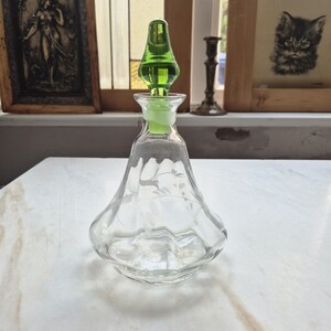 Könnte beinhalten: Eine klare Parfümflasche aus Glas mit einem grünen Verschluss. Die Flasche hat einen ausgestellten Boden und ein geätztes Design. Der Verschluss ist leuchtend grün und hat eine einzigartige, längliche Form. Die Flasche steht auf einer weißen Oberfläche.