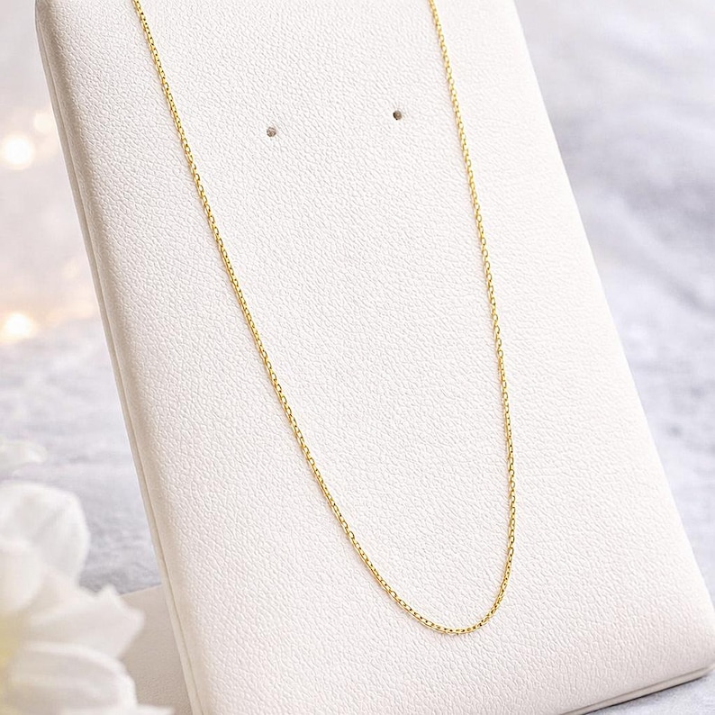 Puede incluir: Un delicado collar de cadena dorada exhibido en un expositor de joyas blanco. La cadena es fina y est&aacute; hecha de peque&ntilde;os eslabones conectados. El fondo est&aacute; suavemente borroso, resaltando la simple elegancia del collar.