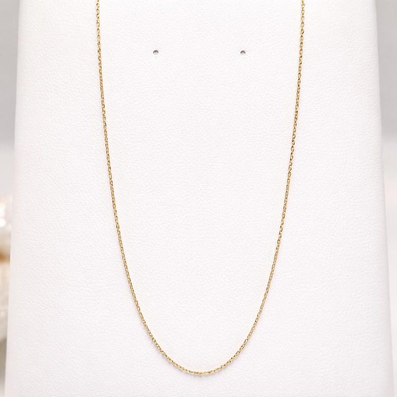 Puede incluir: Un delicado collar de cadena dorada exhibido sobre un fondo blanco. La cadena es fina y parece estar hecha de peque&ntilde;os eslabones interconectados. El collar es una pieza de joyer&iacute;a sencilla y elegante.