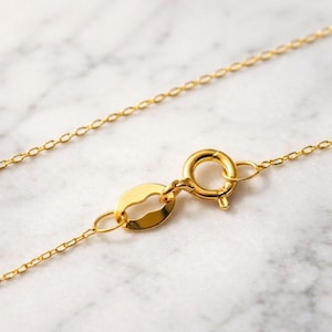 Puede incluir: Primer plano de un collar dorado con una cadena delicada y un cierre de anilla. El collar se muestra sobre una superficie de m&aacute;rmol blanco y gris. Los eslabones de la cadena son peque&ntilde;os y ovalados.
