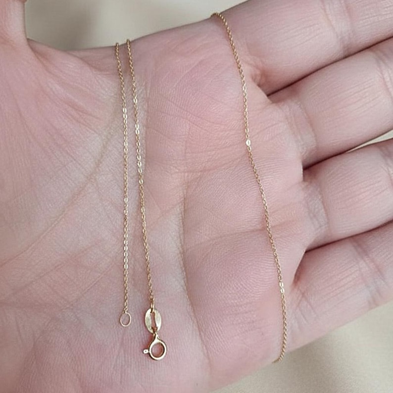 Puede incluir: Un delicado collar de cadena de color dorado con un peque&ntilde;o cierre y un anillo circular. El collar se muestra sobre una superficie de color claro. La cadena es fina y parece estar hecha de peque&ntilde;os eslabones conectados.