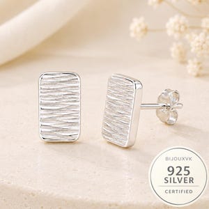 May include: A pair of rectangular silver stud earrings featuring a textured, wavy surface pattern. Each earring has a polished edge and a standard post back with a butterfly clutch. A circular seal in the corner reads BIJOUXVK 925 SILVER CERTIFIED.
