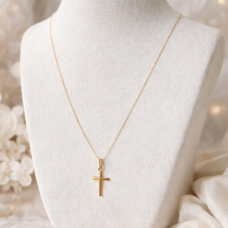 Puede incluir: Un collar dorado con un colgante de cruz. La cruz tiene un dise&ntilde;o sencillo y cl&aacute;sico, y la cadena es fina. El collar se exhibe sobre un busto blanco, con un fondo suave y borroso.