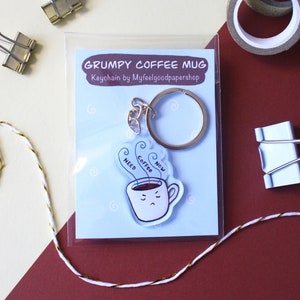 Könnte beinhalten: Ein weißer Kaffeetassen-Schlüsselanhänger mit einem mürrischen Gesicht und dem Text "Need Coffee Now". Der Schlüsselanhänger befindet sich auf einem blauen Hintergrund mit einem silbernen Schlüsselring.