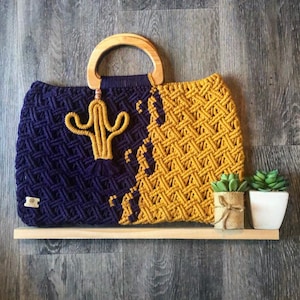 May include: A handmade macrame tote bag with a wooden handle. The bag is divided into two sections: one in navy blue and the other in mustard yellow. A yellow cactus design is attached to the navy side. The bag is displayed on a wooden shelf.