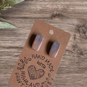 May include: Rectangular stud earrings with a blue and gold metallic finish. The earrings are displayed on a brown card with the text "Hand Made With Highland Craft". The background is a wooden surface.