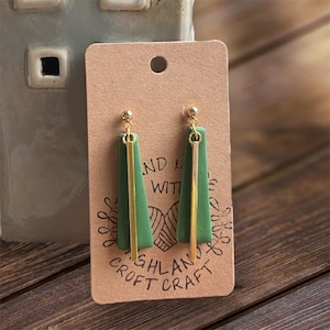 May include: Green and gold dangle earrings on a brown card. The earrings feature a gold ball stud, a green trapezoid shape, and a gold vertical bar. The card has the text "AND WITH" and "HIGHLAND CROFT CRAFT".