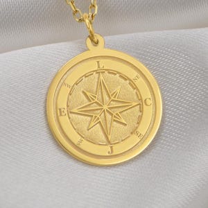May include: A gold-coloured pendant necklace with a compass rose design. The round pendant has a raised border with directional letters and a central starburst. The necklace chain is delicate and gold-toned, resting on a white satin surface.