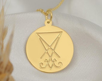 May include: A gold necklace with a round pendant featuring a sigil engraved in the centre. The pendant is on a white fabric background.