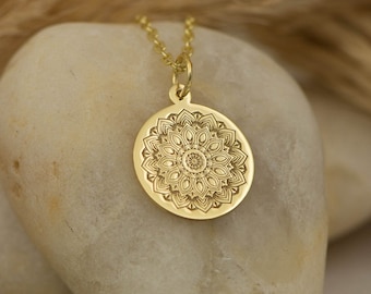 May include: A gold necklace with a round pendant featuring a detailed mandala design. The pendant is on a white rock with a brown background.