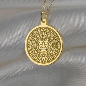 May include: A gold-toned circular pendant necklace featuring an intricate embossed design of a central face surrounded by geometric patterns. The pendant hangs from a delicate gold-toned chain, showcasing a polished metallic finish throughout.