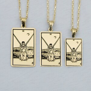 Puede incluir: Tres collares de oro con colgantes que representan la carta del tarot Dos de Espadas. Los colgantes son rectangulares y tienen una ilustración en blanco y negro de una figura que sostiene dos espadas.