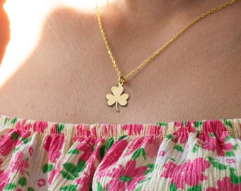 May include: A gold necklace with a four-leaf clover pendant. The necklace is worn by a person wearing a pink and green floral print top.