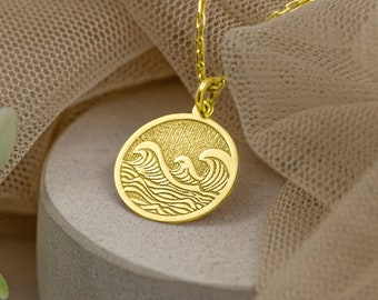 May include: A gold necklace with a round pendant featuring a detailed wave design. The necklace is displayed on a light brown fabric background.