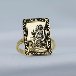 Puede incluir: Anillo de oro con un diseño en negro y oro que presenta una imagen de la Reina de Copas de una carta de tarot.