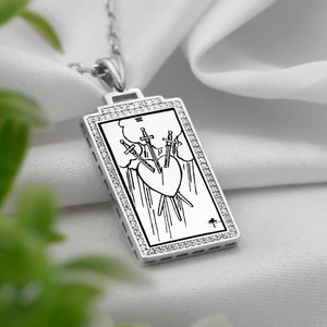 Puede incluir: Un collar de plata con un colgante rectangular que presenta un dise&ntilde;o de carta de tarot. La carta representa el Tres de Espadas, con tres espadas que atraviesan un coraz&oacute;n. El colgante est&aacute; rodeado por un borde de peque&ntilde;os diamantes.