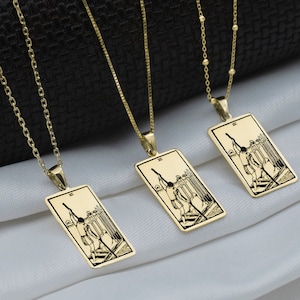 Puede incluir: Cuatro collares de oro con colgantes de cartas del tarot. Cada colgante presenta una ilustración en blanco y negro de una carta del tarot. Las cartas son La Torre, Los Enamorados, El Carro y El Mundo.