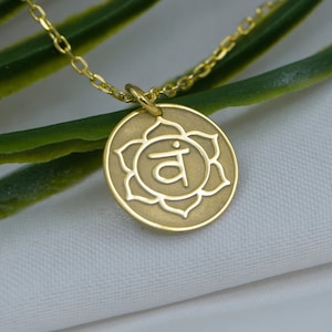 Pode incluir: Colar de ouro com um pingente que representa o símbolo do chakra da garganta, uma flor estilizada com a letra sânscrita 'Vam' no seu interior.