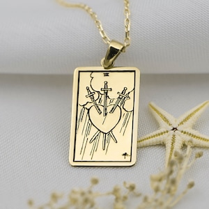 Puede incluir: Collar de oro con un colgante que representa la carta del tarot Tres de Espadas. La carta representa tres espadas que atraviesan un corazón, simbolizando la angustia y la tristeza.