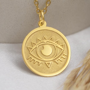 以下が含まれることがあります： 目のデザインが施された丸いペンダントが付いたゴールドネックレス。ペンダントは、スタイリッシュな目と放射状の線で詳細に描かれています。