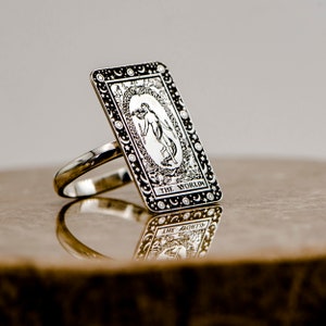 May include: A silver ring with a rectangular design featuring the World tarot card. The card is surrounded by a border of stars and moons. The ring is on a brown surface.